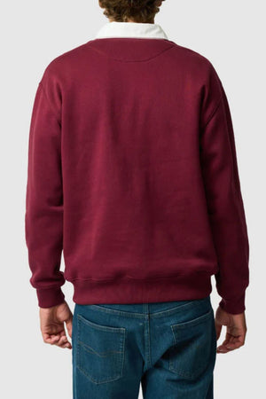 A person is seen from behind wearing a maroon Rodd & Gunn Otago Sweat with a classic rugby collar and blue jeans. The background is plain light gray, and their hands rest relaxed at their sides.