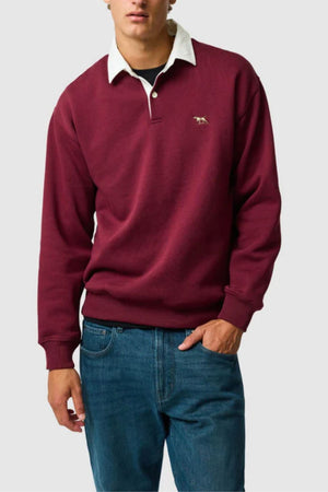 Someone stands against a plain light gray background wearing the Rodd & Gunn Otago Sweat in maroon, featuring a white rugby collar and small embroidered logo, paired with blue jeans.