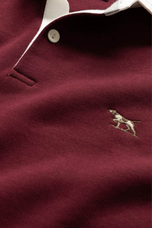 A close-up of the Rodd & Gunn Otago Sweat, a maroon polo with a rugby collar, white buttons and trim, and an embroidered dog logo on the chest.