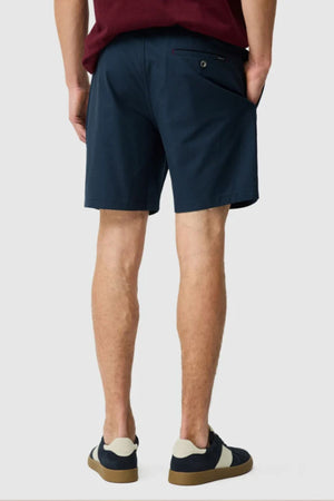 A person stands with hands in pockets, wearing Rodd & Gunn Resort Shorts in dark blue cotton with an elastic waist, paired with a burgundy shirt and navy sneakers featuring beige and brown soles, set against a plain light gray background.