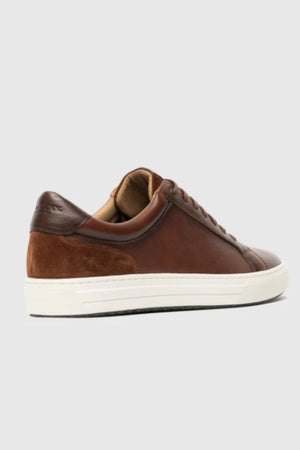 The Rodd & Gunn Transit Sneaker is a brown leather and suede low-top with a white rubber sole, shown from the back-left against a plain light gray background.