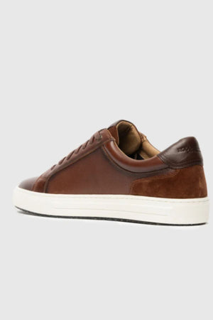 The Rodd & Gunn Transit Sneaker, a brown leather shoe with suede accents, white rubber sole, and brown laces, is displayed from the rear side against a plain light gray background.