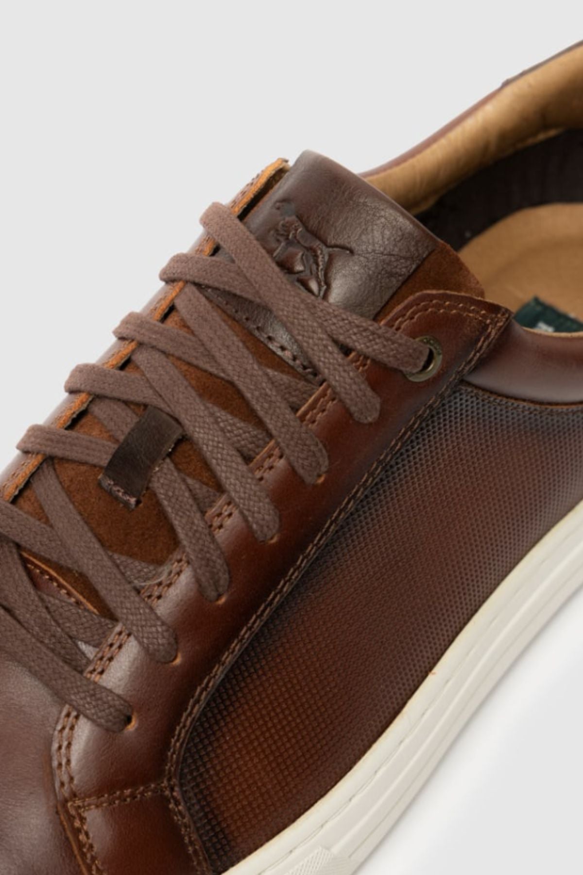 Close-up of the Rodd & Gunn Transit Sneaker: brown leather with textured side panel, brown laces, and white rubber sole. Embossed logo on tongue and visible stitching showcase its premium craftsmanship.
