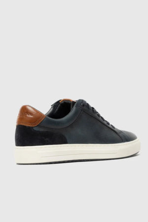 The Rodd & Gunn Transit Sneaker showcases a black leather design, white sole, black laces, and a brown heel accent, displayed on a light gray background.