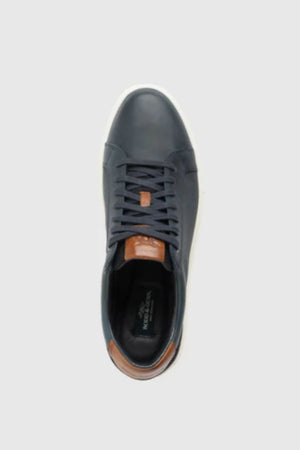 The Rodd & Gunn Transit Sneaker features navy blue leather with brown leather accents on the tongue and heel, black laces, and a white sole, displayed from above on a light gray background.