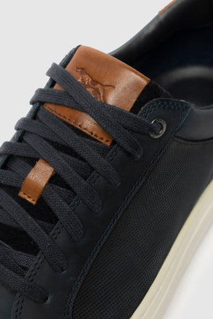 A close-up of the Rodd & Gunn Transit Sneaker, a black leather shoe with dark laces, a white sole, and brown leather on the tongue featuring an embossed animal design.