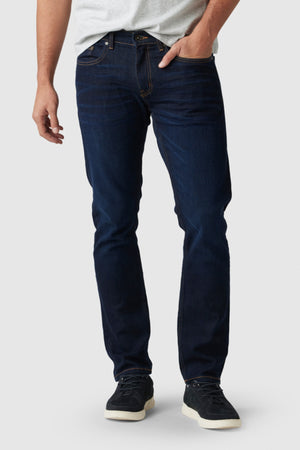 A man in a light gray t-shirt, Rodd & Gunn Fanshawe Straight Jeans, and black sneakers with white soles stands with one hand in his pocket against a plain light background.
