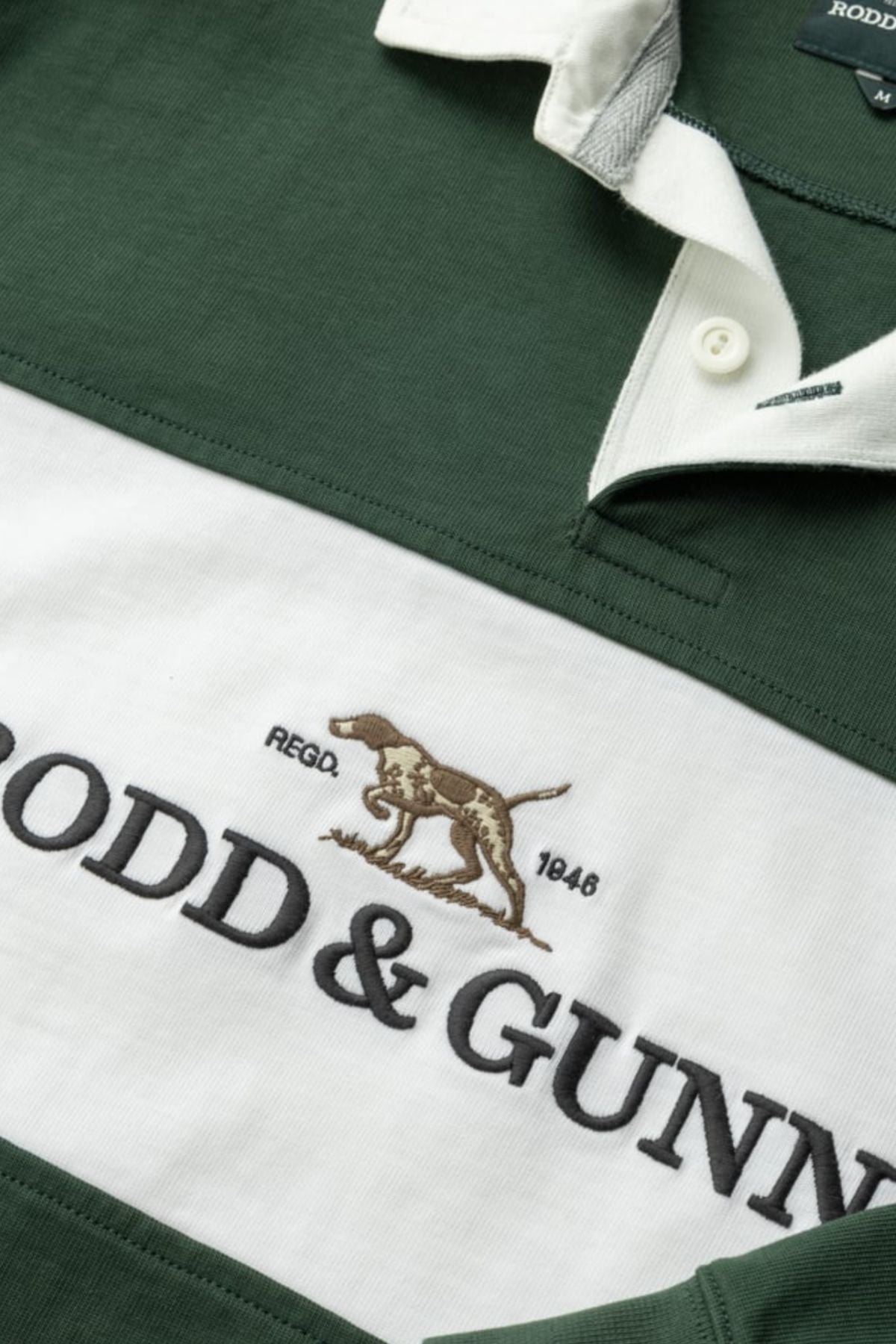 Close-up of the Rodd & Gunn Foresters Rugby shirt in green and white 100% cotton jersey, showing part of the collar, buttons, “RODD & GUNN” text, an embroidered dog logo, and “REGO. 1848” on the white stripe.
