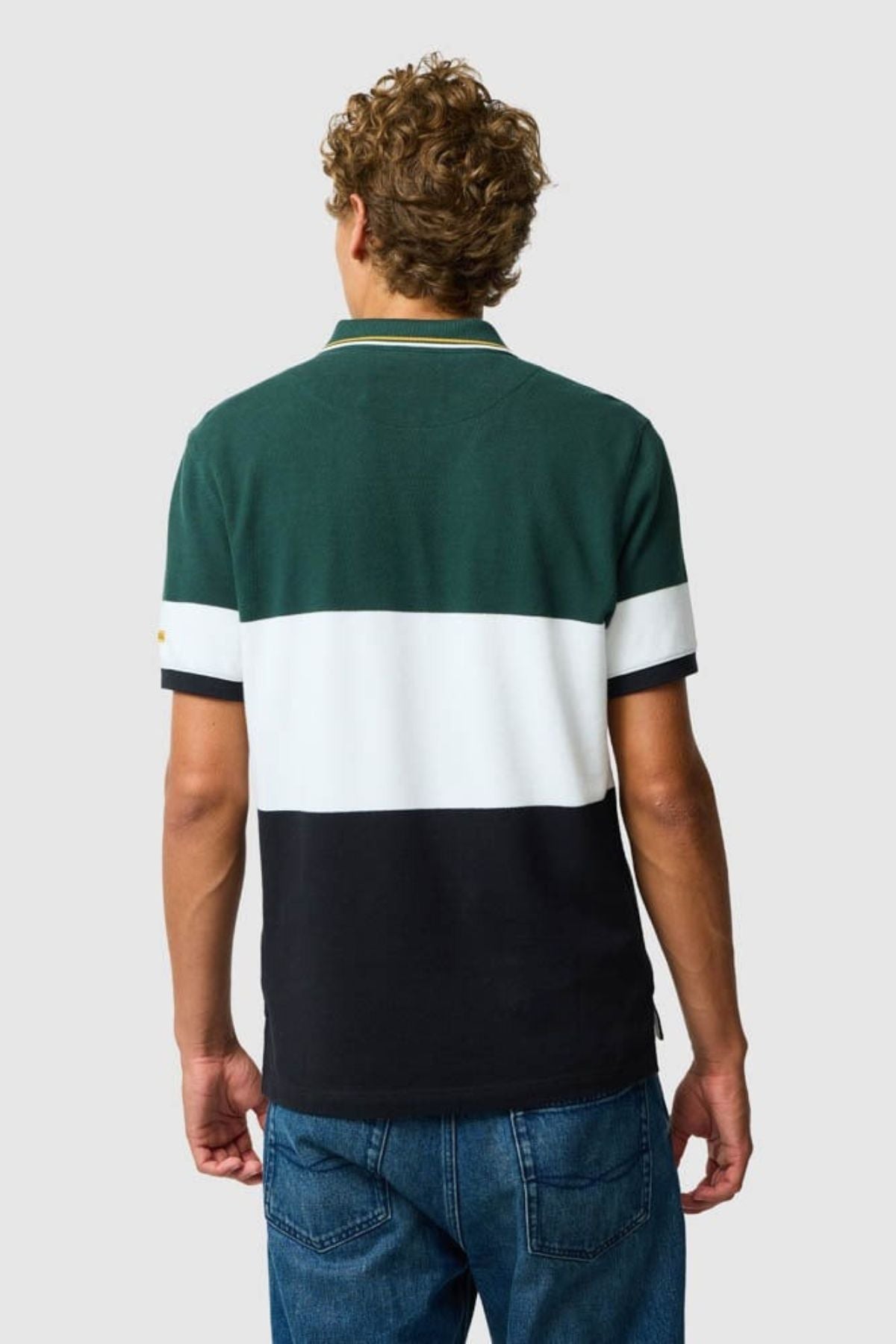 A person with curly hair, dressed in a Rodd & Gunn Homebay Polo made from Australian Good Earth Cotton in green, white, and black color-blocked stripes and blue jeans, stands with their back to the camera against a plain background.