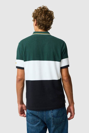 A person with curly hair, dressed in a Rodd & Gunn Homebay Polo made from Australian Good Earth Cotton in green, white, and black color-blocked stripes and blue jeans, stands with their back to the camera against a plain background.