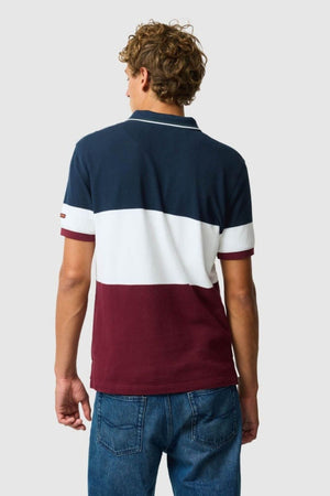 Wearing the Rodd & Gunn Homebay Polo—a breathable, sustainable short-sleeved pique knit with navy, white, and maroon color-blocking—and blue jeans, a curly-haired person stands with their back to a plain light background.