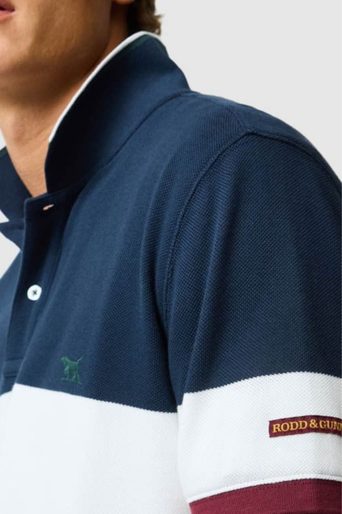 A person wears the Rodd & Gunn Homebay Polo—navy blue and white, collared, with a small green dog logo on the chest and "RODD & GUNN" patch on the sleeve—crafted from breathable pique knit. Only their lower face and upper torso are visible.