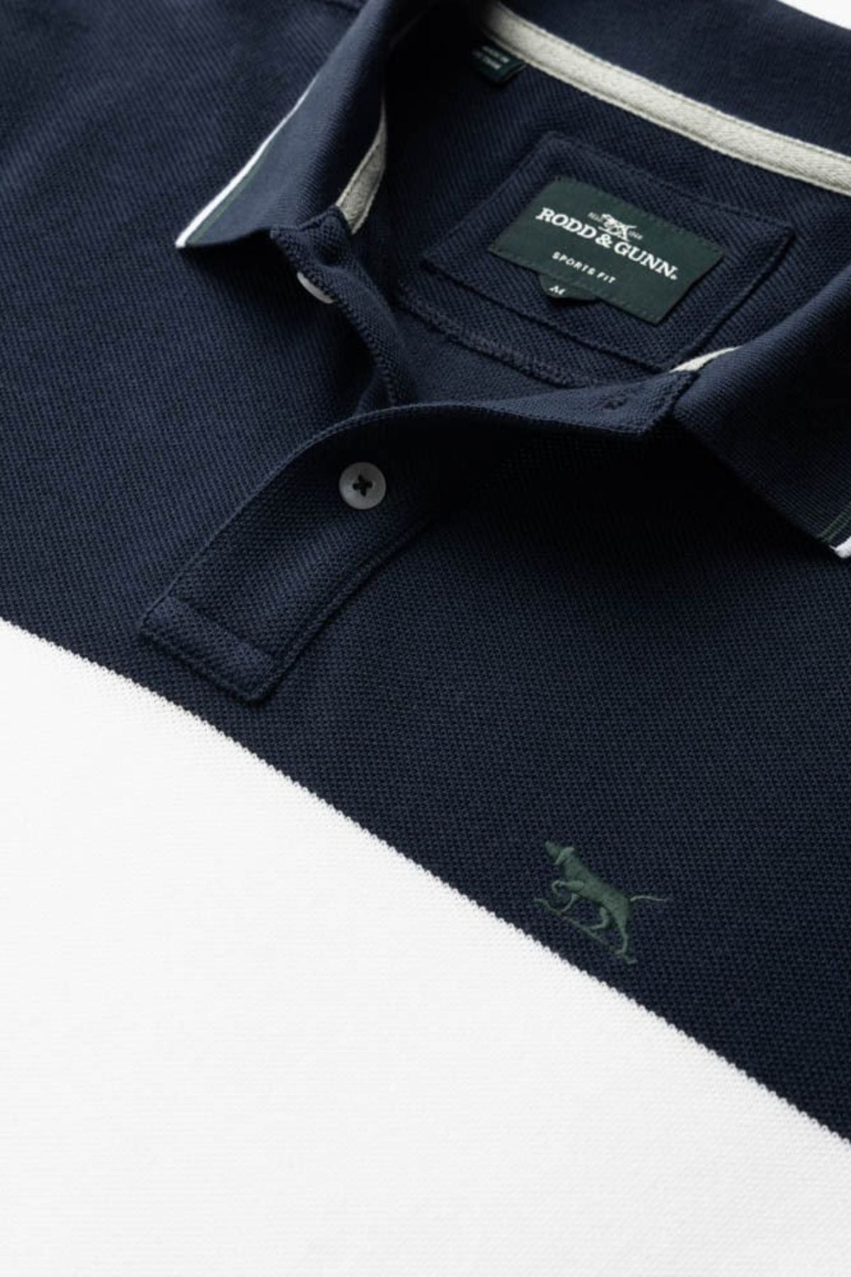 Close-up of the Rodd & Gunn Homebay Polo in navy blue and white color-blocking, crafted from breathable pique knit with a collar, buttons, and embroidered dog logo. The tag shows "Rodd & Gunn, Family Run.