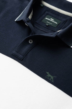 Close-up of the Rodd & Gunn Homebay Polo in navy blue and white color-blocking, crafted from breathable pique knit with a collar, buttons, and embroidered dog logo. The tag shows "Rodd & Gunn, Family Run.