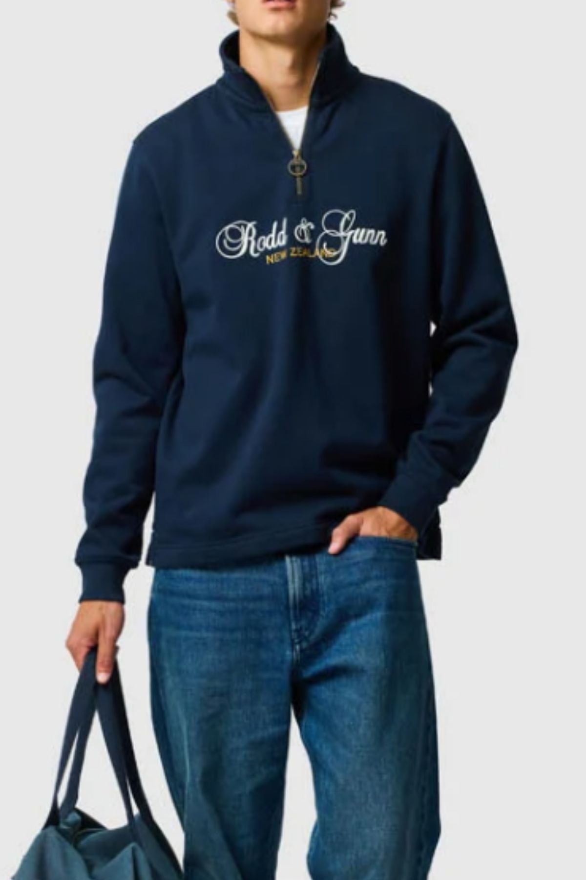 Wearing the Rodd & Gunn Jubilee Sweat, a navy quarter zip with "Rodd & Gunn New Zealand," and blue jeans, they hold a matching navy bag in their left hand against a plain, light-colored background.