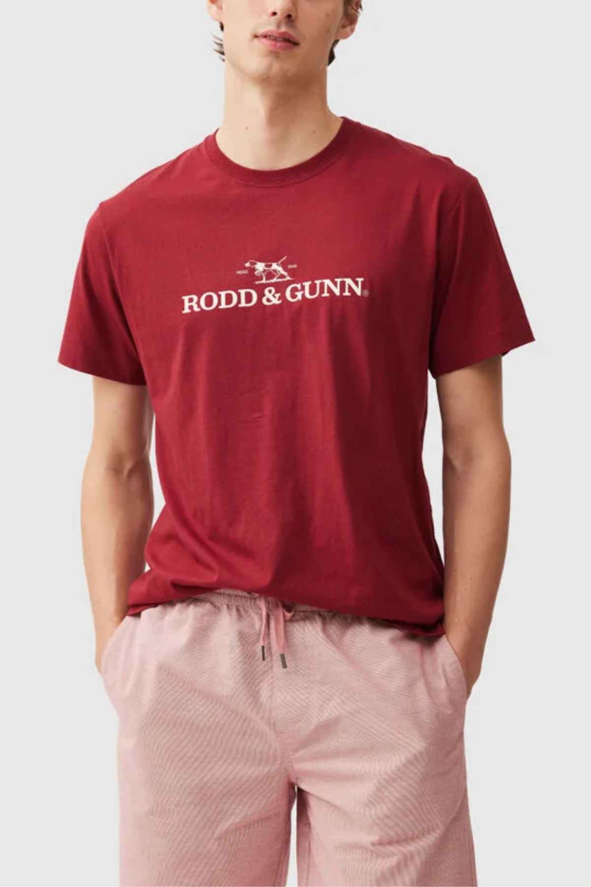 A person is wearing a Rodd & Gunn Logo T-Shirt made from sustainably farmed cotton with a small dog graphic, paired with light pink shorts, hands in pockets against a plain light gray background.