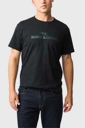 The individual is dressed in a Rodd & Gunn Logo T-Shirt made from sustainably sourced cotton, set against a simple background. The sports fit black shirt complements their dark jeans, with their arms relaxed at their sides.