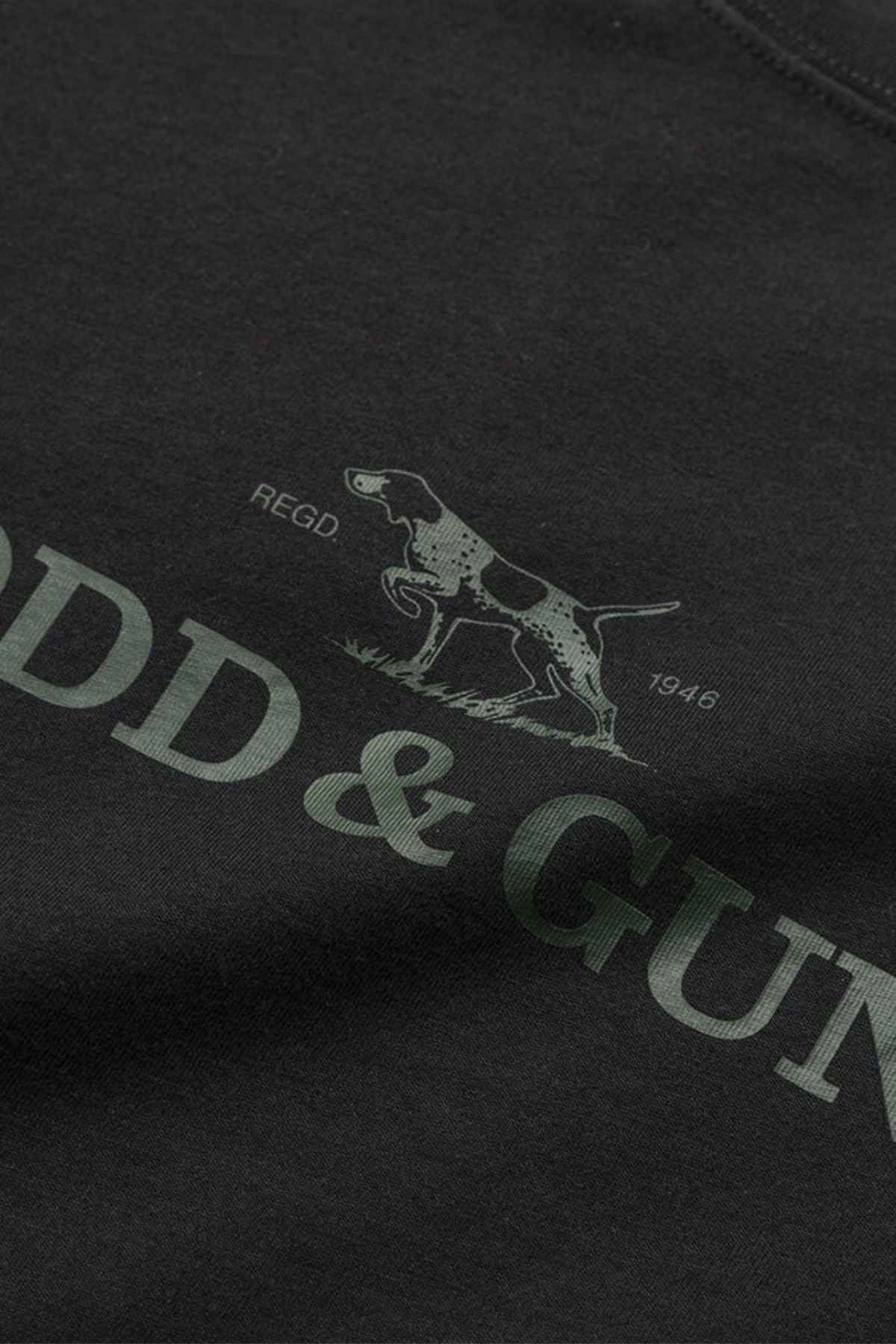 Close-up of a black fabric features a green design, including an illustration of a dog and the text "REGD 1946." The words "ODD & GUN" are partially visible beneath the image, suggesting it's the renowned Rodd & Gunn Logo T-Shirt made from sustainably farmed cotton.