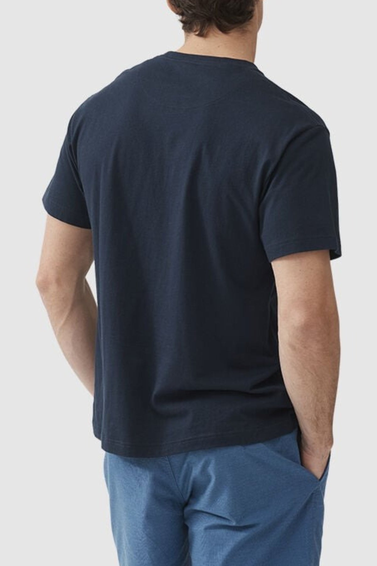 Rear view of a person standing with hands in pockets, donning a dark navy Rodd & Gunn Logo T-Shirt and light blue pants. The individual has short, wavy hair and a light skin tone. The background is plain white.