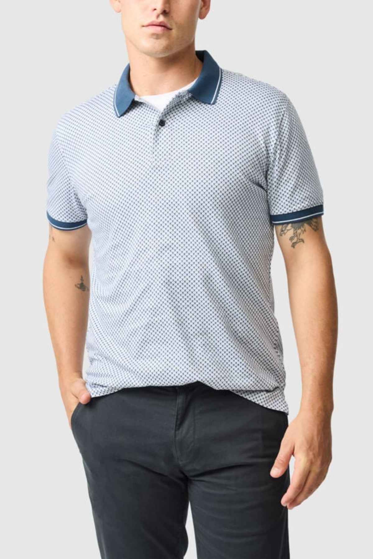 A person dressed in a Rodd & Gunn Lynden Sports Fit Polo, featuring a blue pattern and crafted from luxurious Turkish cotton, pairs it with dark pants while standing with one hand in their pocket against a plain background.