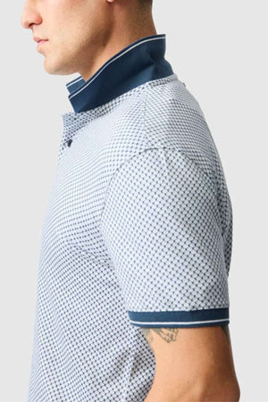 A person wearing the Rodd & Gunn Lynden Sports Fit Polo in light blue, accented with a dark blue collar and cuffs. Crafted from luxurious Turkish cotton, this shirt showcases small geometric shapes, enhancing their style. The collar is slightly turned up, revealing a tattoo on their left arm.