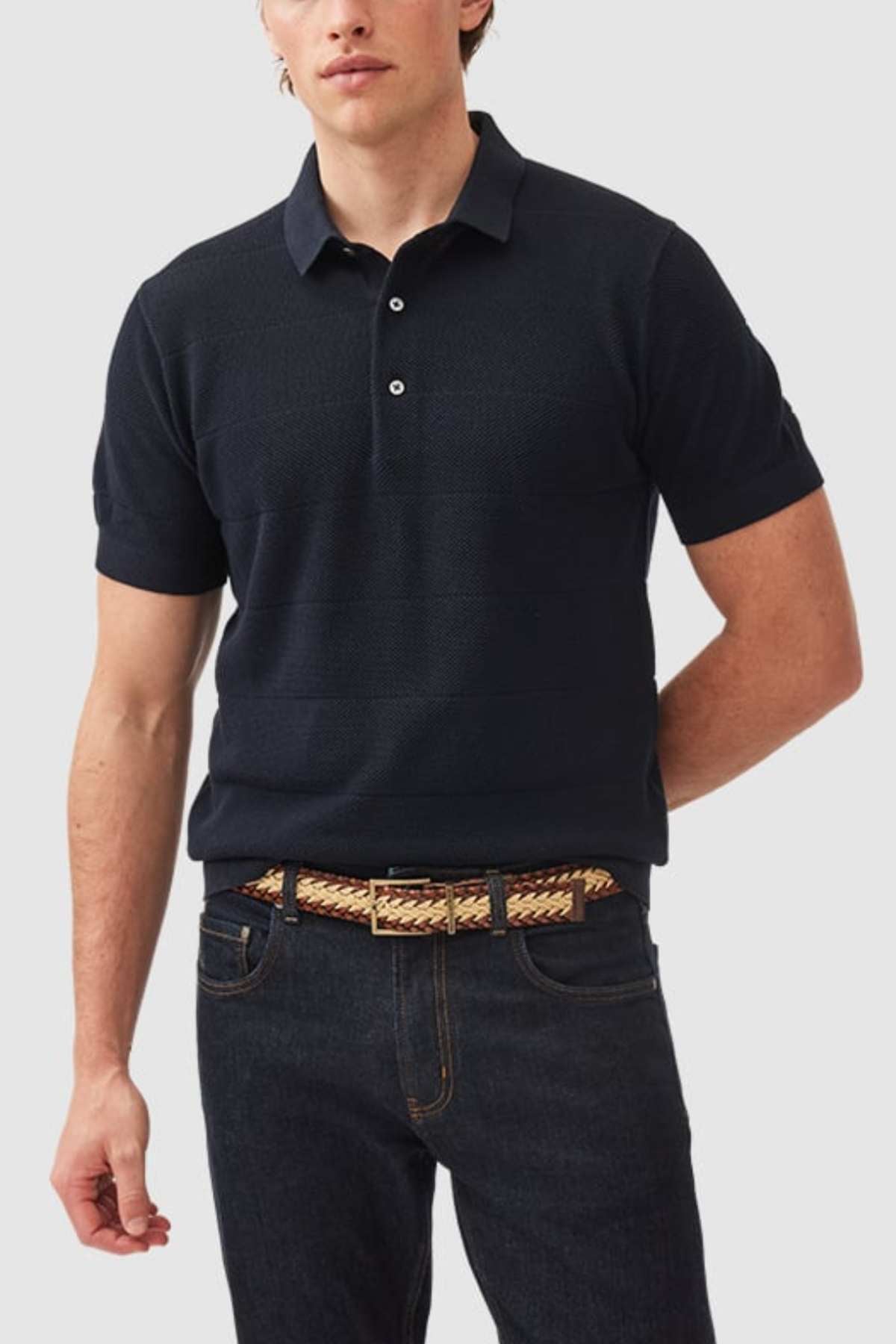 A man wearing a Rodd & Gunn Masons Flat Knit short-sleeve polo shirt in dark navy and dark blue jeans stands against a plain white background. His left arm is behind his back, and he is wearing a woven beige and brown belt. The polo shirt features a collar and three buttons.