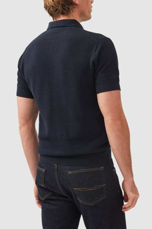 A man with short, light brown hair is facing away from the camera, wearing a dark navy Rodd & Gunn Masons Flat Knit short-sleeve polo shirt and dark blue jeans. The plain, light-colored background emphasizes the clothing.