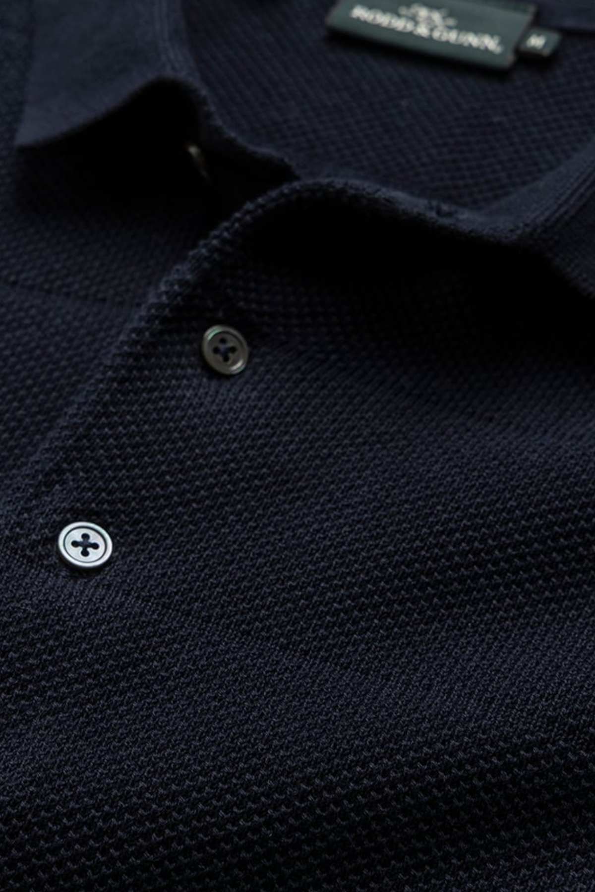 Close-up shot of the Rodd & Gunn Masons Flat Knit polo shirt in black, showcasing its textured knit fabric and collar with two white buttons. The brand label is clearly stitched into the back of the collar area, with the intricate knit pattern and fine stitching details prominently visible.