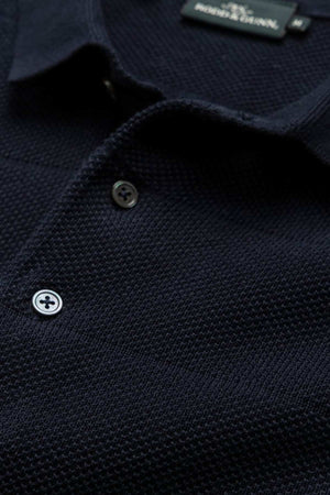 Close-up shot of the Rodd & Gunn Masons Flat Knit polo shirt in black, showcasing its textured knit fabric and collar with two white buttons. The brand label is clearly stitched into the back of the collar area, with the intricate knit pattern and fine stitching details prominently visible.