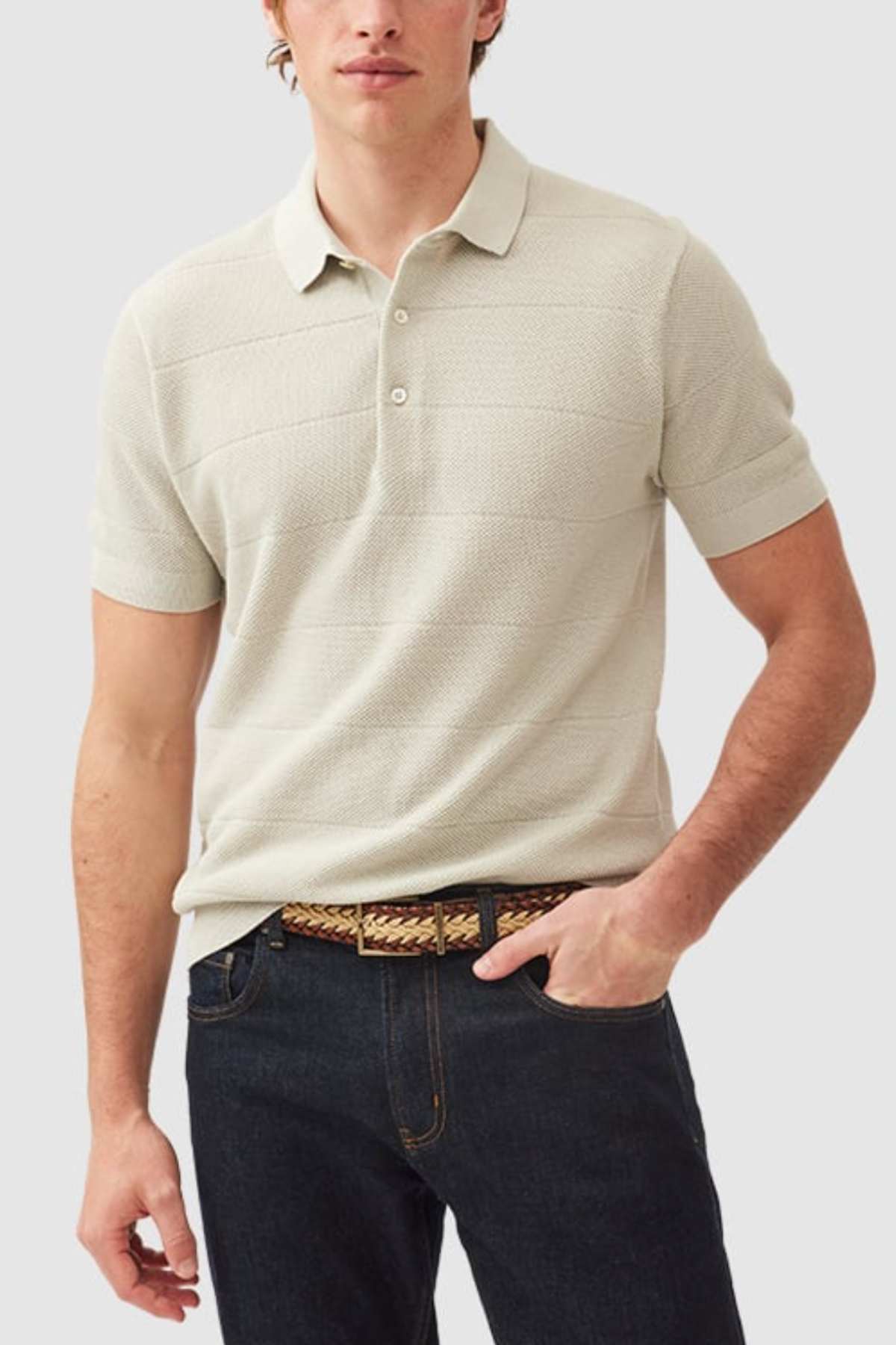 A person wearing a Rodd & Gunn Masons Flat Knit light beige cotton polo with a classic pique stitch and dark jeans stands with one hand in their pocket, sporting a brown woven belt against the plain gray background.