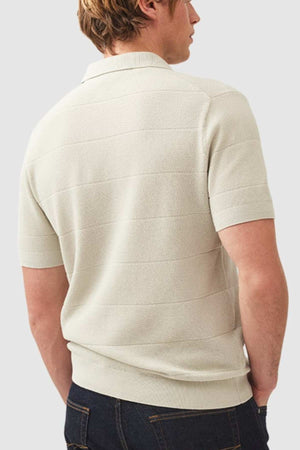 A light brown-haired person wearing the Rodd & Gunn Masons Flat Knit, a light beige short-sleeve polo with classic pique stitch, faces away. The shirt is paired with dark jeans against a plain gray background.