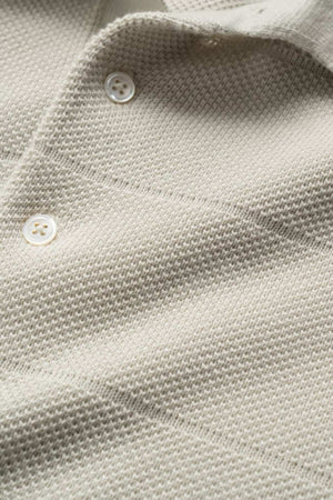Close-up of a textured beige fabric with a classic pique stitch pattern and three white buttons. The soft, lightweight material resembles the Rodd & Gunn Masons Flat Knit, ideal for clothing such as shirts or cardigans.