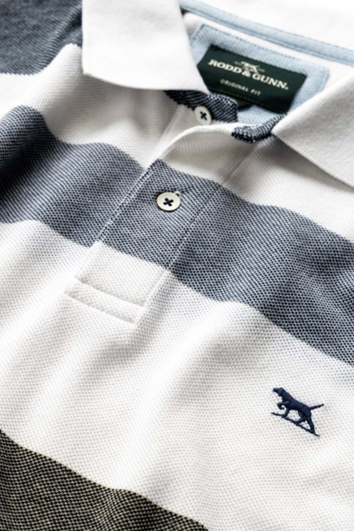 Close-up of the Rodd & Gunn Milton Polo made from USA cotton, with wide white and gray stripes. It has a two-button collar and a small embroidered dog logo on the right chest.