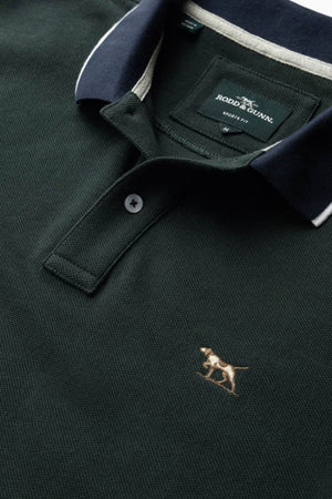 A close-up of the Rodd & Gunn New Lynn Polo: a dark green men's sports-fit polo featuring a navy collar, white trim, and an embroidered dog logo. Made from Australian Good Earth Cotton; label reads "Rodd & Gunn, Sports Fit.