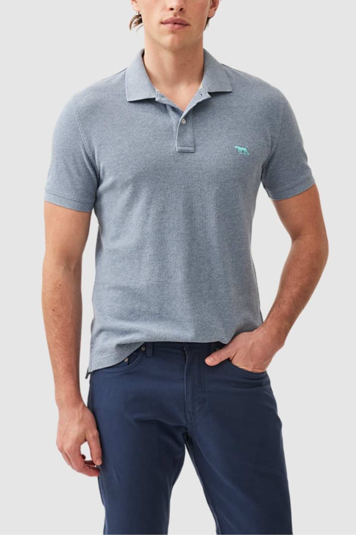 A man is wearing a Rodd & Gunn The Gunn Polo in light blue and dark blue pants. The polo shirt features a small embroidered logo, and the man has one hand in his pocket. The background is plain and light-colored.