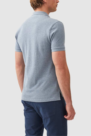 A person is shown from the back wearing the Rodd & Gunn The Gunn Polo in light blue and navy blue pants. Their head is slightly turned to the right, and they have short, light brown hair. The background is a simple, plain light grey.