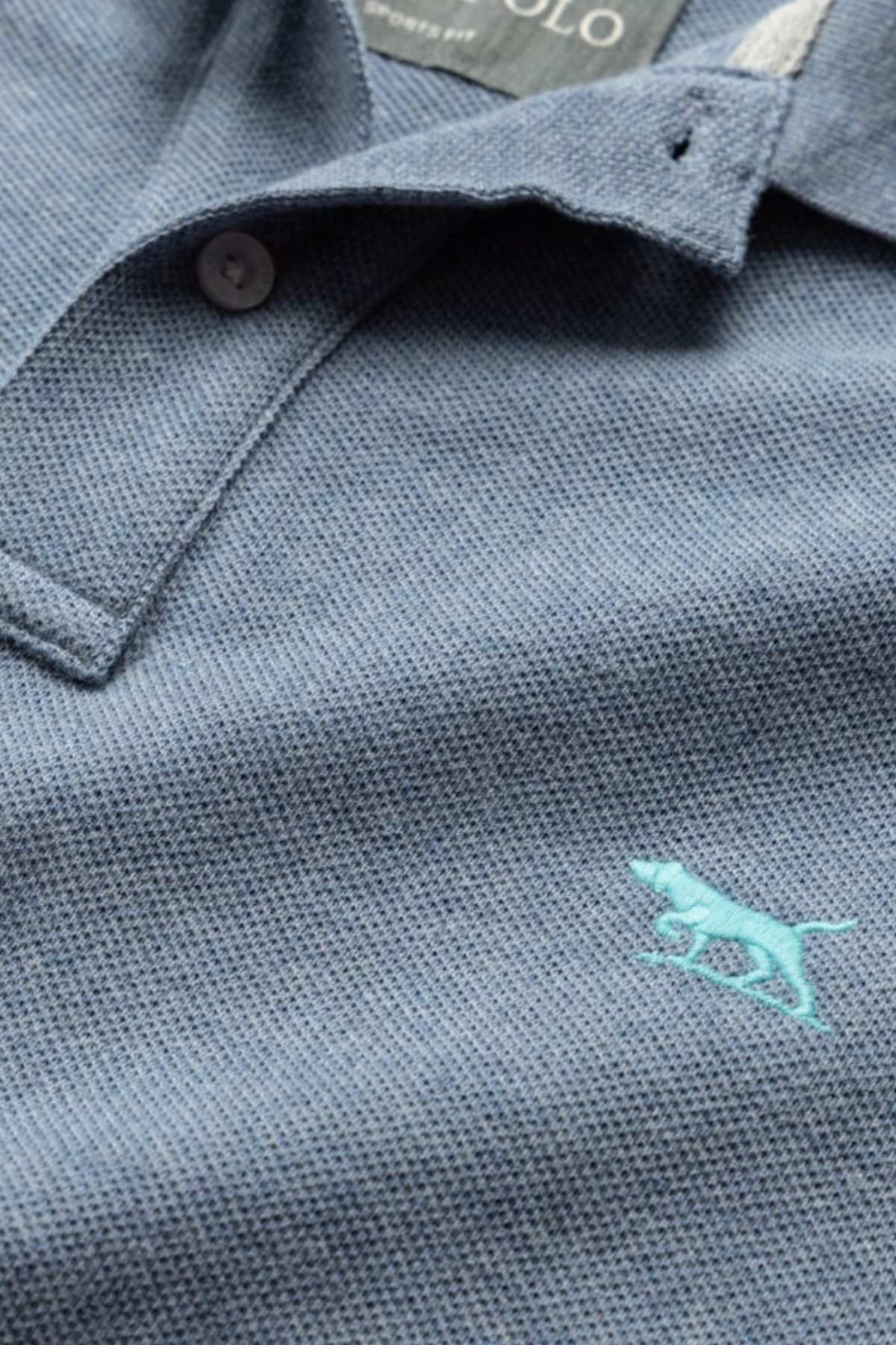Close-up of a blue textured Rodd & Gunn The Gunn Polo shirt with a buttoned collar. Visible on the shirt is an embroidered light blue dog logo near the bottom of the picture. The shirt tag indicates the brand name but is not fully visible.