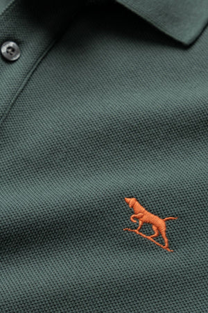 Close-up of the Rodd & Gunn The Gunn Polo, a dark green textured fabric casual shirt featuring an embroidered orange dog logo on the left side. Crafted from premium Australian Cotton, this modern fit polo shirt has a button placket visible at the top.