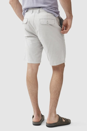 A person stands facing away from the camera, wearing light gray Rodd & Gunn The Gunn Shorts, featuring back pockets. They pair it with a tucked-in light purple shirt and dark brown sandals. The background is plain and light-colored.