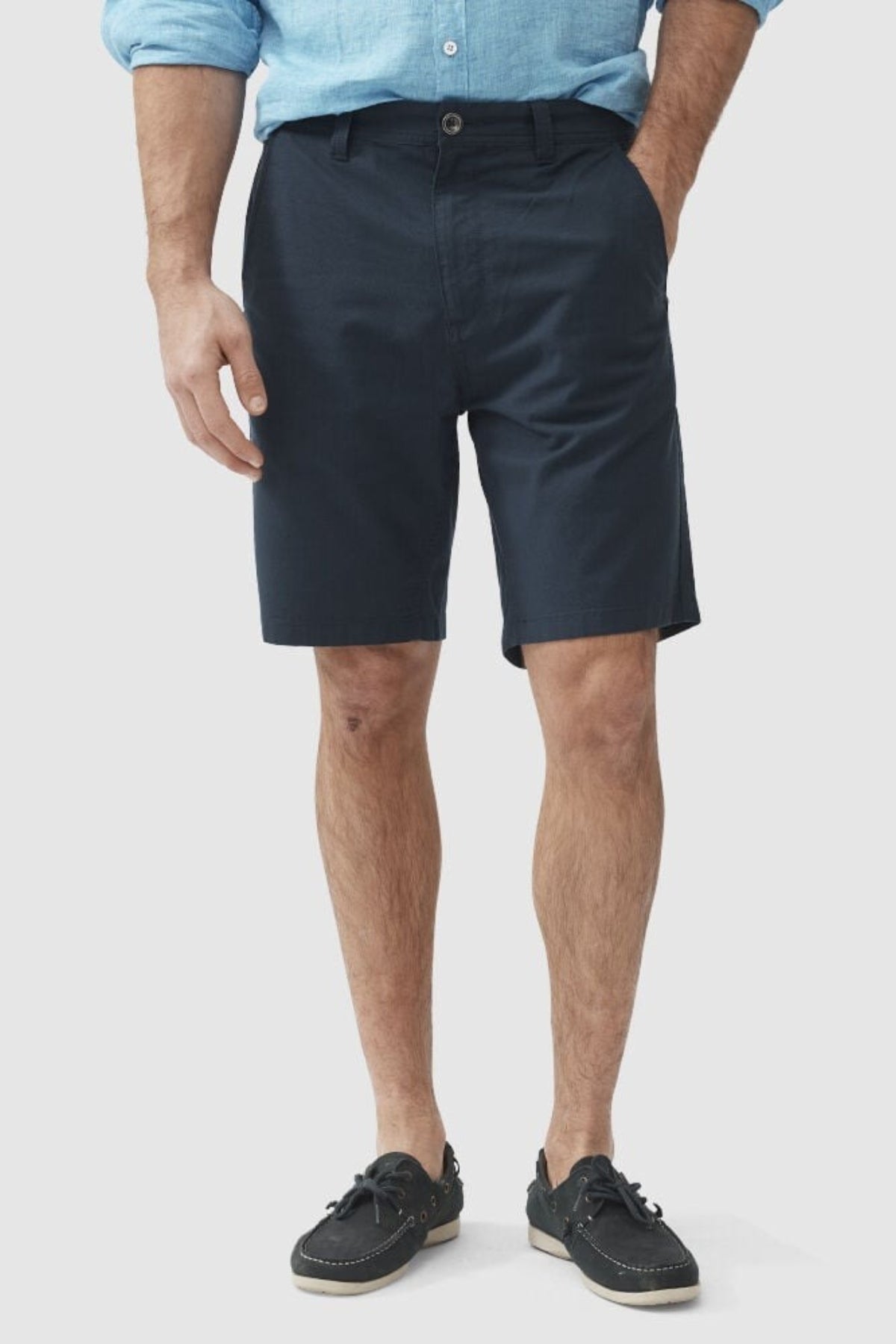 An individual is wearing a light blue long-sleeve shirt made from a cotton elastane blend, tucked into Rodd & Gunn's The Gunn Short in classic navy. They are also sporting black boat shoes with white soles. The background is plain white.