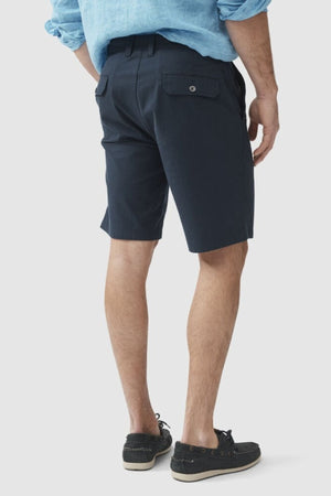 Rear view of a person wearing a blue collared shirt, Rodd & Gunn The Gunn Short in dark knee-length classic fit with buttoned pockets made from a cotton elastane blend, and black boat shoes. The person's hands are relaxed at their sides, and they are standing against a plain white background.