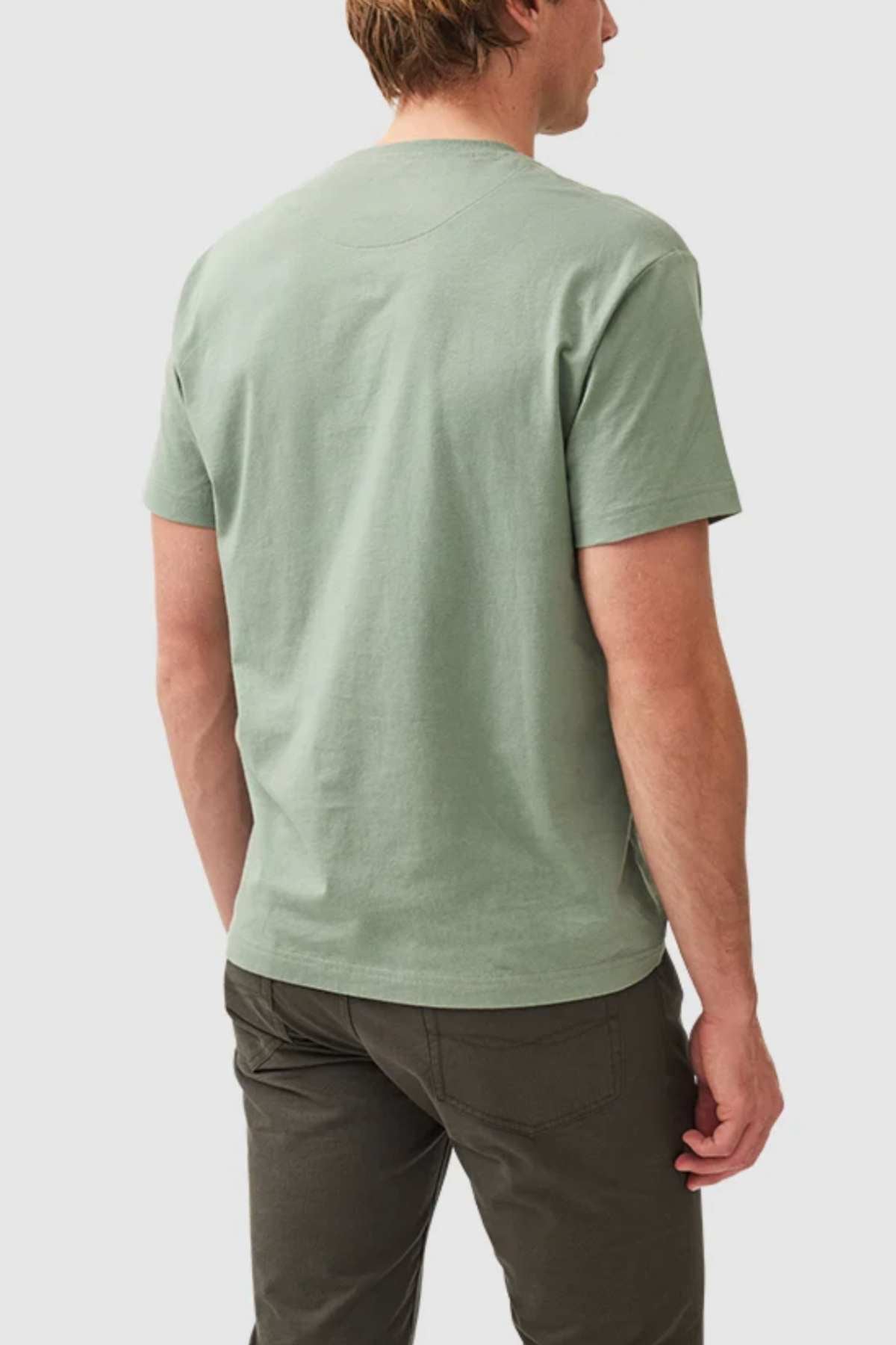 A person with light hair is seen from behind, wearing a sage green Rodd & Gunn The Gunn T-Shirt and dark green pants. The background is plain white.
