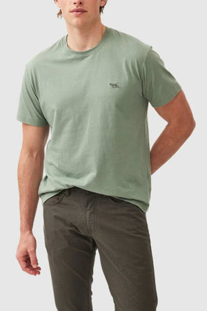 A person wearing the Rodd & Gunn The Gunn T-Shirt in light green, featuring a small embroidered logo on the chest, and dark pants stands against a plain background. Their left hand is in their pocket, and they have light skin and medium-length, slightly wavy brown hair.
