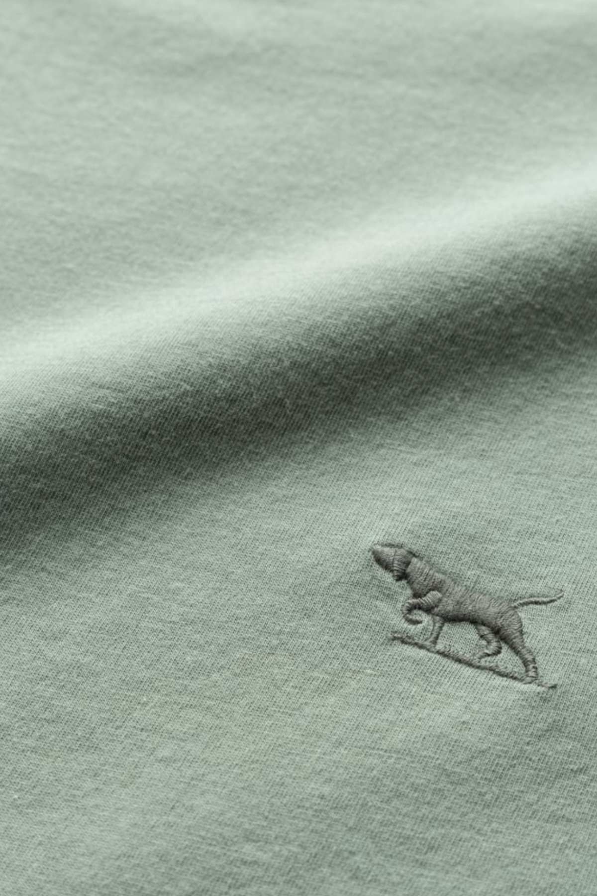 A close-up image of the light green fabric of the Rodd & Gunn The Gunn T-Shirt, featuring a small embroidered logo of a dog. The dog, which appears to be walking, is stitched in a slightly darker shade of green. The fabric boasts a soft and smooth texture.