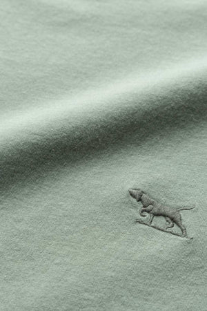 A close-up image of the light green fabric of the Rodd & Gunn The Gunn T-Shirt, featuring a small embroidered logo of a dog. The dog, which appears to be walking, is stitched in a slightly darker shade of green. The fabric boasts a soft and smooth texture.