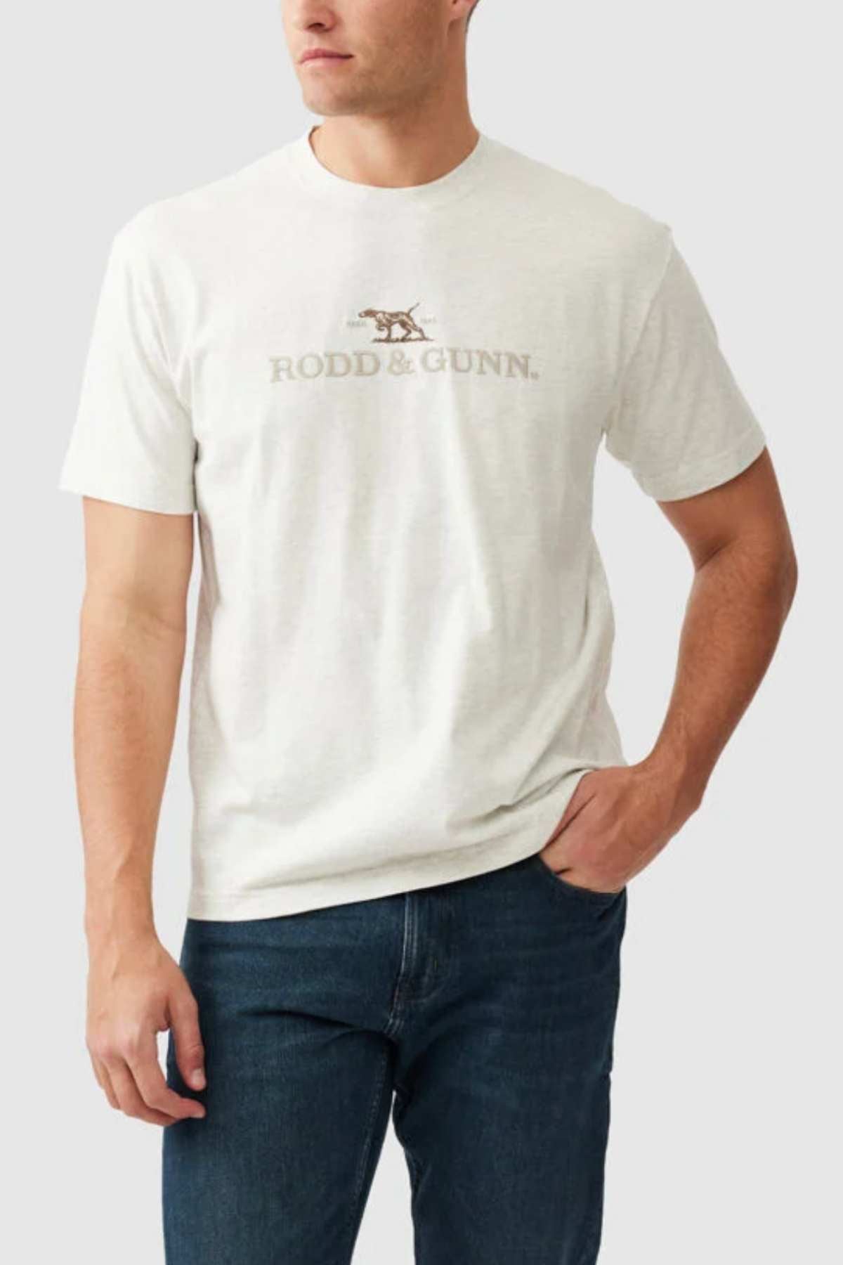 A person is wearing the "Rodd & Gunn The Peaks Logo Tee," a light-colored T-shirt featuring the "RODD & GUNN" logo and a small graphic of a hunting dog, crafted from Australian Good Earth Cotton. The T-shirt is paired with dark jeans, set against a plain white background.