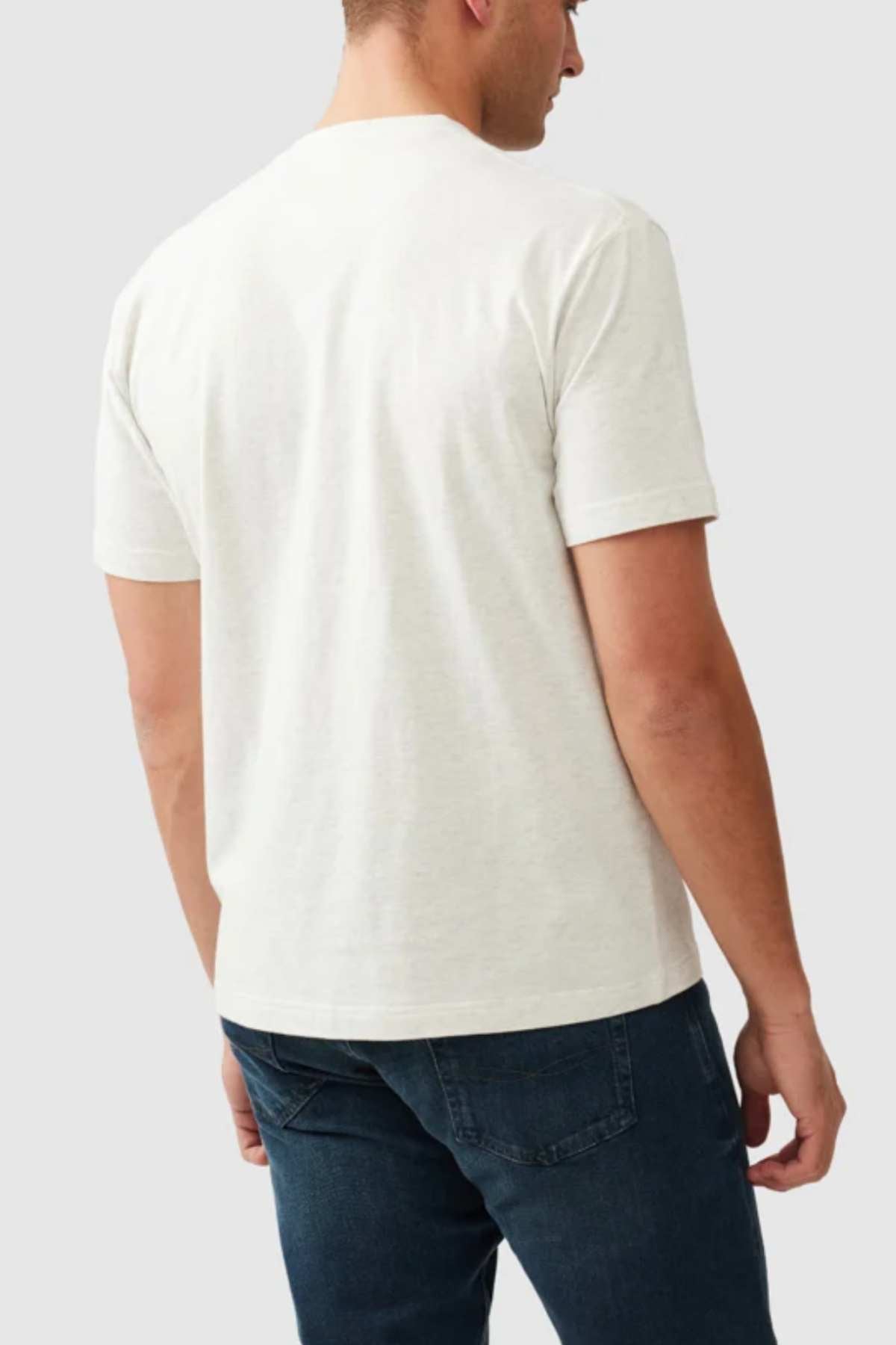 A man stands facing away, wearing the Rodd & Gunn The Peaks Logo Tee, a simple white short-sleeve T-shirt, paired with dark blue jeans. The background is plain and light-colored, emphasizing the casual outfit. A subtle Rodd & Gunn logo adds a touch of refinement to the ensemble.