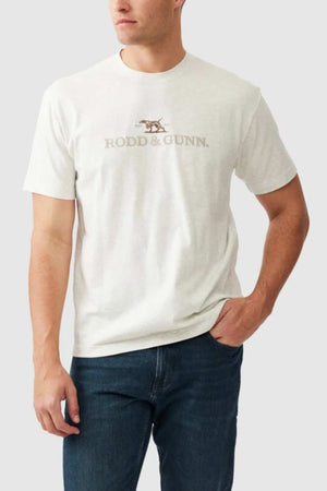A person is wearing the "Rodd & Gunn The Peaks Logo Tee," a light-colored T-shirt featuring the "RODD & GUNN" logo and a small graphic of a hunting dog, crafted from Australian Good Earth Cotton. The T-shirt is paired with dark jeans, set against a plain white background.