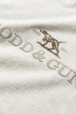 Close-up image of light-colored fabric with an embroidered design on the Rodd & Gunn The Peaks Logo Tee. The embroidery features the Rodd & Gunn logo along with a graphic of a dog standing and looking forward, accompanied by the text "REG'D 1916." This design also highlights the synergy of FibreTrace technology woven into Australian Good Earth Cotton.