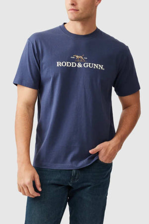 A person stands against a plain background wearing the Rodd & Gunn The Peaks Logo Tee in navy blue, featuring the "RODD & GUNN" logo and an image of a dog in a running stance on the front. The individual has one hand in their pocket and is dressed casually in jeans.